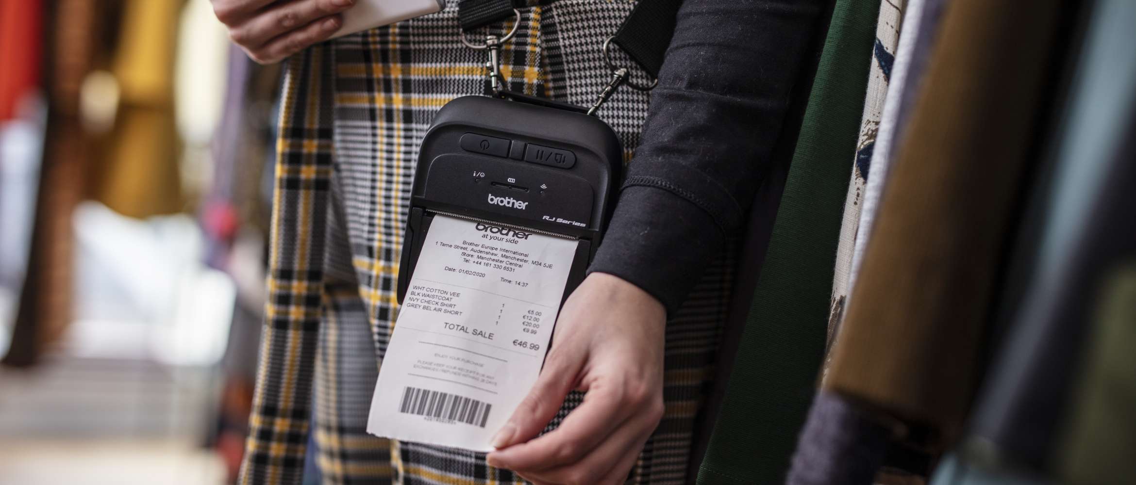 A Brother handheld labelling and receipt printer is worn on-body of a store assistant of a retail clothing shop. It is printing out a customer receipt. 